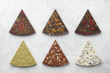 Triangle or pizza slice shaped chocolate pieces with diverse kinds of toppings on white textured background, top viewの写真素材