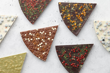 Triangle or pizza slice shaped chocolate pieces with diverse kinds of toppings on white textured background, top viewの写真素材