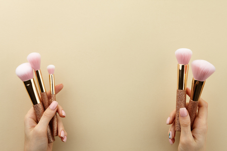 Womans hands holding golden glossy make up brushes with pink bristles on beige background, close up view, copy spaceの写真素材