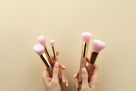 Womans hands holding golden glossy make up brushes with pink bristles on beige background, close up view, copy spaceの写真素材