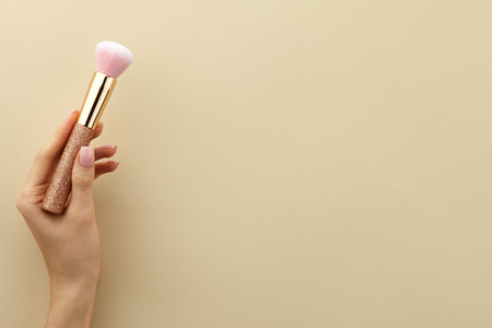 Womans hand holding golden glossy make up brush with pink bristles on beige background, close up view, copy spaceの写真素材