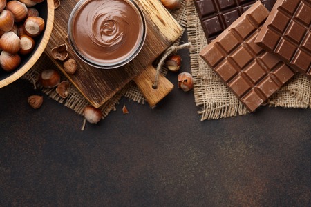 Chocolate spread or nougat cream with hazelnuts in glass jar, bowl of nuts and chocolate bars on brown countertop, copy spaceの写真素材