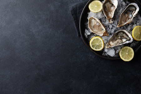 Fresh opened oysters in a plate with ice and lemon on black textured background, top viewの写真素材