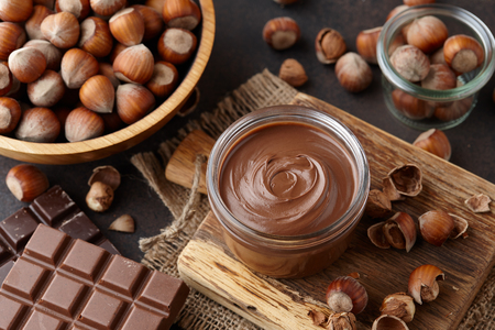 Chocolate spread or nougat cream with hazelnuts in glass jar, bowl of nuts and chocolate bars on brown countertopの写真素材