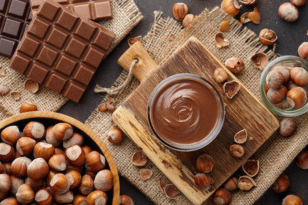 Chocolate spread or nougat cream with hazelnuts in glass jar, bowl of nuts and chocolate bars on brown countertopの写真素材
