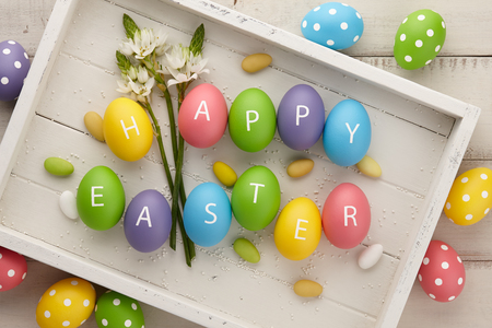 Colorful Easter eggs with letters arranged in Happy Easter greeting and spring flowers on white wooden planks. Holiday concept.の写真素材
