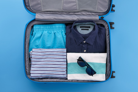 Open travel suitcase with mens shirts, shorts, tshirts and other summer clothes on blue background, top viewの写真素材