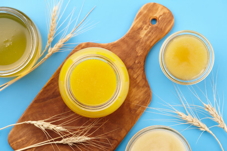 Different kinds of honey in glass jars and wheat ears on wooden cutting board on blue background, copy spaceの写真素材