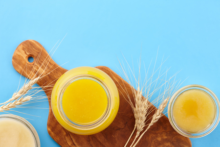 Different kinds of honey in glass jars and wheat ears on wooden cutting board on blue background, copy spaceの写真素材