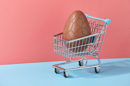 Easter chocolate egg with bow in shopping basket on trendy pink and blue background, creative holiday conceptの写真素材