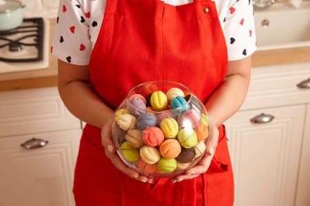 Woman in red apron holding glass bowl with colorful cookies in the kitchen, close up viewの写真素材