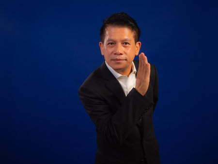 Portrait of smart middle-aged asian confident successful businessman wearing suit whispering something on blue background in studio, looking attractive and positive professional leadershipの写真素材