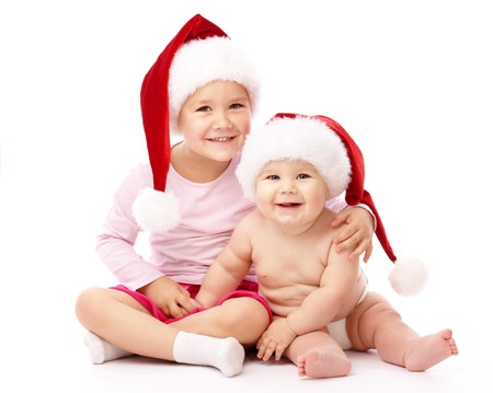 Girl hugs her little brother, both wearing red Christmas caps and smile, isolated over whiteの写真素材