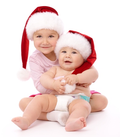 Girl hugs her little brother, both wearing red Christmas caps and smile, isolated over whiteの写真素材