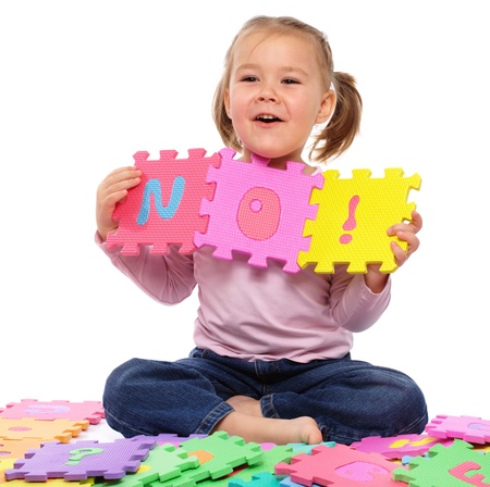 Cute little girl is playing with alphabet, put together word No while sitting on floor, isolated over whiteの写真素材