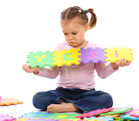 Sad little girl is playing with alphabet, put together word Yes while sitting on floor, isolated over whiteの写真素材