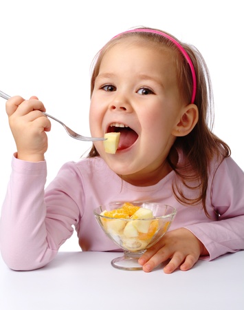 Cute little girl eats fruit salad using fork, isolated over whiteの写真素材