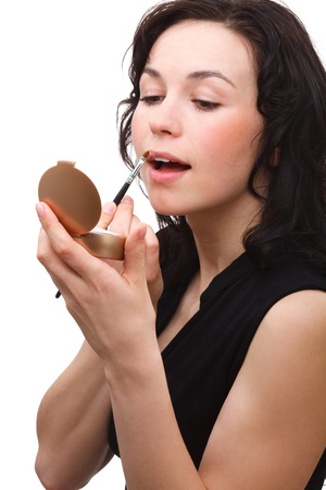 Young woman is applying lipgloss using brush while looking at herself in mirror, isolated over whiteの写真素材