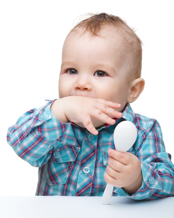 Cute little boy holding spoon and biting his finger while sitting at table, isolated over whiteの写真素材