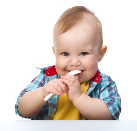 Cute little boy is biting a spoon while sitting at table, isolated over white