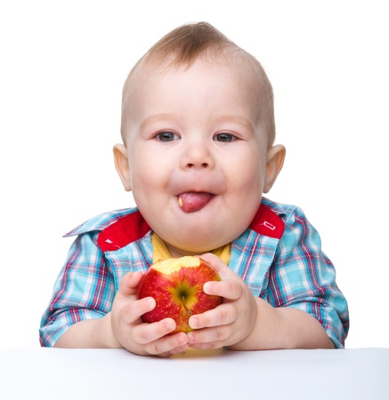 Cute little child is eating red apple while sitting at table, isolated over whiteの写真素材