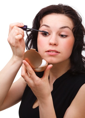 Young woman is applying mascara while looking at herself in mirror, isolated over whiteの写真素材