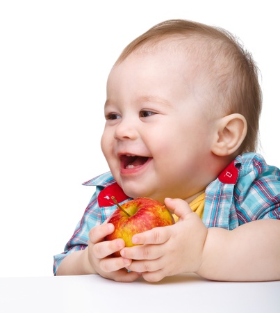 Cute little child is eating red apple and smile while sitting at table, isolated over whiteの写真素材