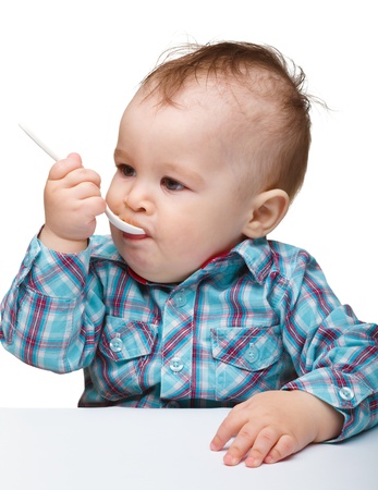 Cute little boy is biting a spoon while sitting at table, isolated over whiteの写真素材