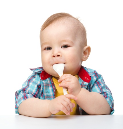 Cute little boy is biting a spoon while sitting at table, isolated over whiteの写真素材