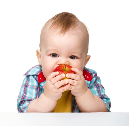 Cute little child is eating red apple while sitting at table, isolated over whiteの写真素材