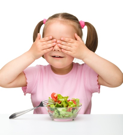 Cute little girl closed her eyes by hands while eating vegetable salad, isolated over whiteの写真素材