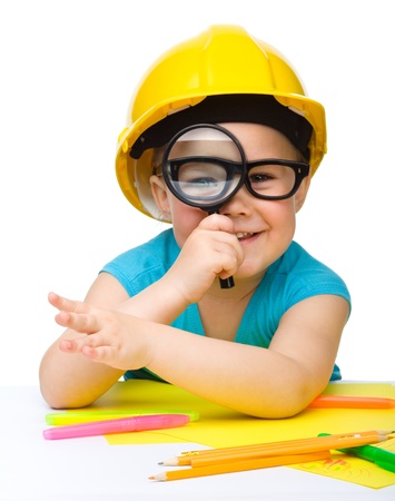 Cute little girl is looking through magnifier wearing hard hat, isolated over whiteの写真素材