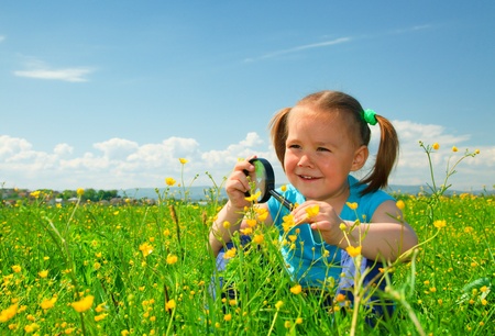 Little girl is playing on green meadow and examining field flowers using magnifying glassの写真素材