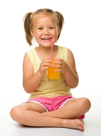 Cute little girl drinks orange juice using drinking straw while sitting on the floor, isolated over whiteの写真素材