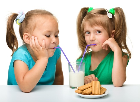 Two Little girls are drinking milk from one glass using straw, isolated over whiteの写真素材