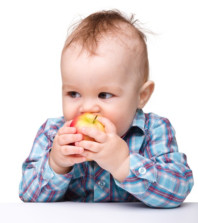 Cute little child is eating red apple while sitting at table, isolated over whiteの写真素材