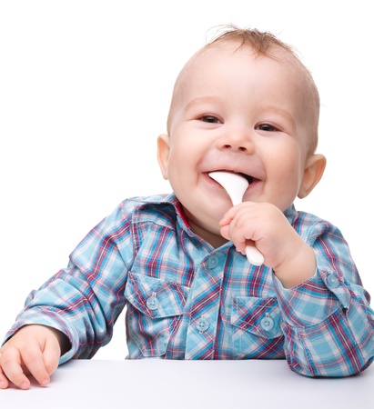 Cute little boy is biting spoon while sitting at table, isolated over whiteの写真素材