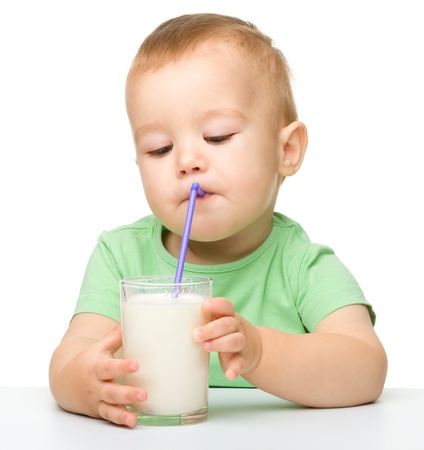 Cute little boy is drinking milk using straw while sitting at table, isolated over whiteの写真素材