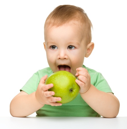 Cute little child is eating green apple while sitting at table, isolated over whiteの写真素材