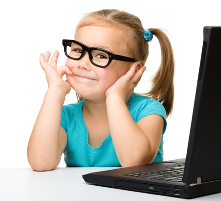 Cute little girl is sitting at table with her black laptop and wearing glasses, isolated over whiteの写真素材