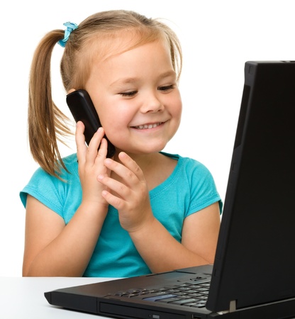 Cute little girl is sitting at table with her black laptop and talking to a cell phone, isolated over whiteの写真素材