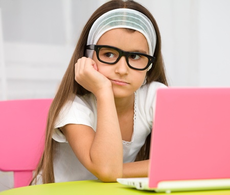 Cute little girl is sitting at table with her pink laptop and wearing glassesの写真素材