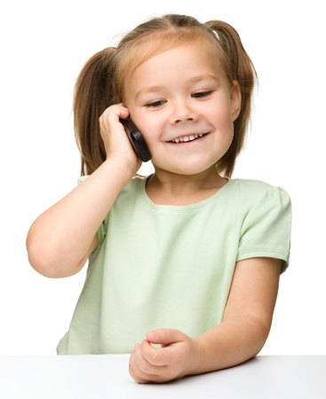 Cute little girl is talking on cell phone while sitting at table, isolated over whiteの写真素材
