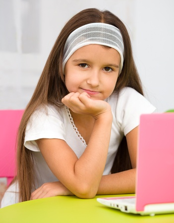 Cute little girl is sitting at table with her pink laptopの写真素材