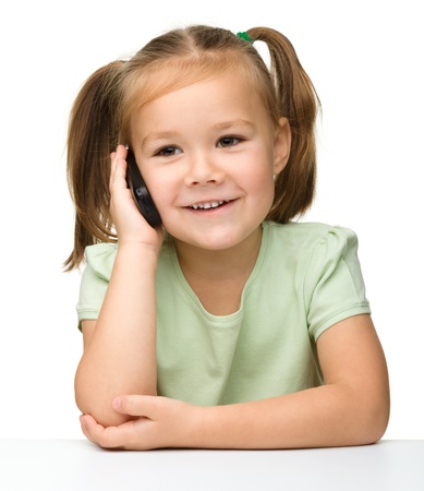 Cute little girl is talking on cell phone while sitting at table, isolated over whiteの写真素材