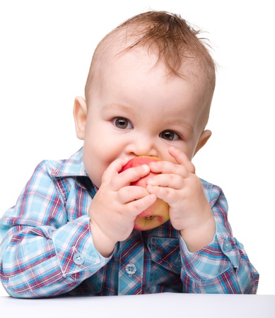 Cute little child is biting red apple and smile while sitting at table, isolated over whiteの写真素材