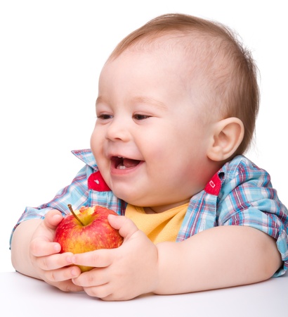 Cute little child is eating red apple and smile while sitting at table, isolated over whiteの写真素材