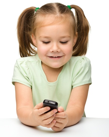 Cute little girl is playing with cell phone while sitting at table, isolated over whiteの写真素材