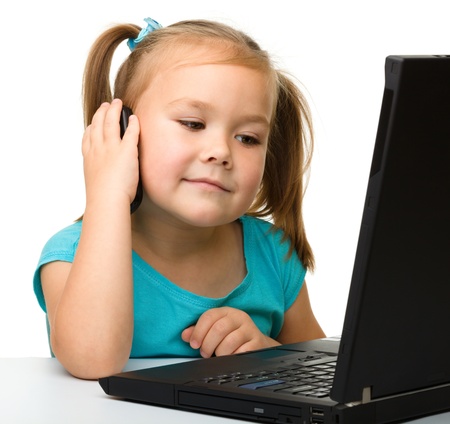 Cute little girl is sitting at table with her black laptop and talking to a cell phone, isolated over whiteの写真素材