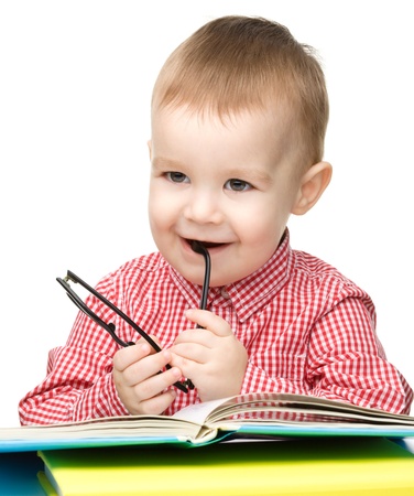 Cute little child play with book and biting glasses while sitting at table, isolated over whiteの写真素材